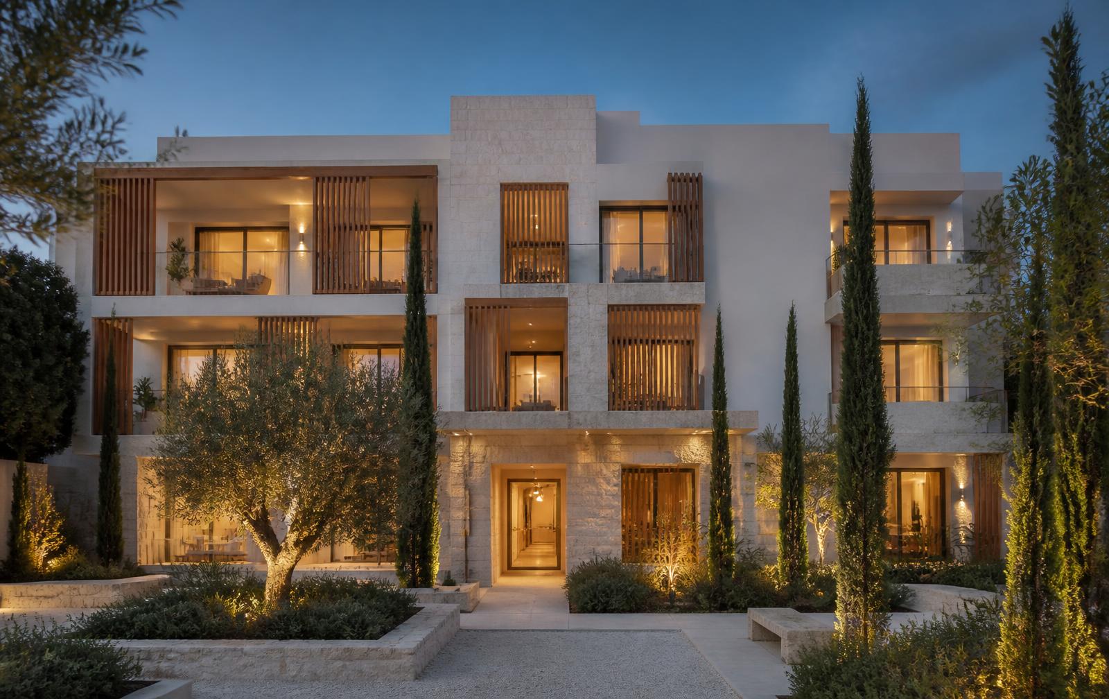 Arvora Collection I at dusk: three-storey white and limestone apartment building with oak balcony screens and a lit courtyard of olive and cypress trees